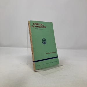 May include: A light green book titled "Spiritual Experiences" by Swami Sivananda. The book has red text and a circular emblem on the cover. The book is propped up on a clear stand.