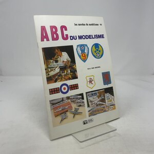 Pode incluir: Um livro intitulado "ABC du Modelisme" com o texto "les mordus du modélisme 10". A capa apresenta imagens de modelos de aviões, uma pessoa trabalhando em um modelo e vários emblemas. O livro é branco com texto rosa e azul.
