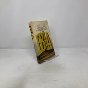 Puede incluir: Un libro de tapa dura titulado "A Revelation in Archaeology" con grandes letras amarillas que deletrean "EBA". El libro tiene una cubierta beige con texto y está apoyado en un soporte de plástico transparente.