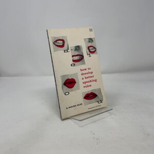 May include: A vintage book titled "how to develop a better speaking voice" by Marjorie Heller. The cover features a cream background with red lips and the letters "a, e, i, o, u". The book is propped up on a clear plastic stand.