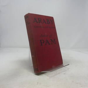 Pode incluir: Um livro vintage de capa dura vermelho intitulado "Araby" de Baroness Von Hutten, com "Author of PAM" impresso na capa. O livro tem letras pretas e é exibido em um suporte transparente.
