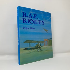 Può includere: Un libro blu con il titolo "R.A.F. KENLEY" e l'autore "Peter Flint" sulla copertina. La copertina presenta un dipinto di un biplano e un hangar sullo sfondo.