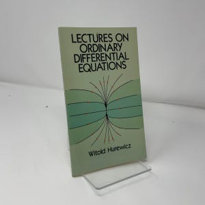May include: A book titled "Lectures on Ordinary Differential Equations" by Witold Hurewicz. The cover is light green with black text and a graphic of curved lines and red dots. The book is propped up on a clear plastic stand.