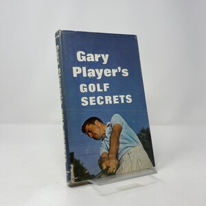 May include: A vintage book titled "Gary Player's Golf Secrets" with a blue cover. The cover features white text and an image of a golfer in action. The book is propped up on a clear plastic stand.