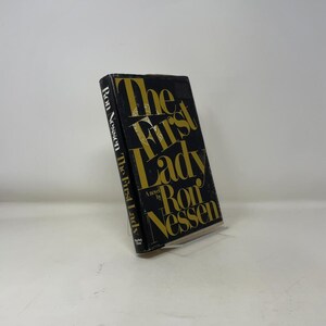 May include: A hardcover book titled "The First Lady" by Ron Nessen. The book has a black cover with gold lettering. The title is prominently displayed, and the author's name is also visible. The book is slightly angled, showcasing its cover design.