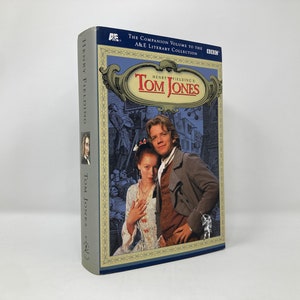 Puede incluir: Una cubierta de libro azul y dorada con el título "Henry Fielding's Tom Jones" y una imagen de dos personas con ropa de época. El libro forma parte de la colección literaria A&E.