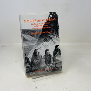 Pode incluir: Um livro intitulado "MY LIFE AS AN INDIAN" com texto laranja e preto. A capa apresenta uma fotografia a preto e branco de nativos americanos. O subtítulo é "The Story of a Red Woman and a White Man in the Lodges of the Blackfeet."