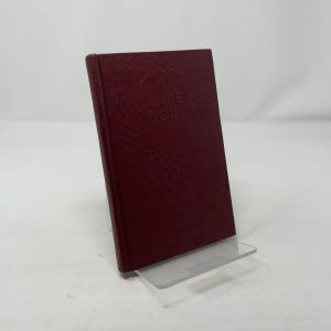 Puede incluir: Un libro de color burdeos con el título "I Dare You" en relieve en la portada. El libro tiene una superficie texturizada y se muestra en un soporte de acrílico transparente. El lomo del libro es visible.