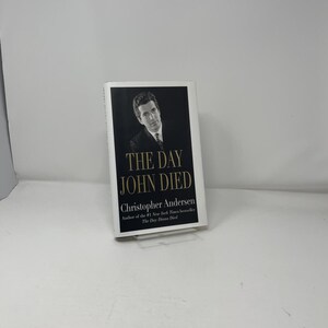 May include: A hardcover book titled "THE DAY JOHN DIED" by Christopher Andersen. The book cover features a black and white image of a man and the title in large white letters. The author's name and book title are also printed on the cover.