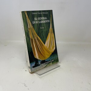 May include: A hardcover book titled "EL GENERAL EN SU LABERINTO" by Gabriel García Márquez. The cover features a yellow hammock against a green background. The book is propped up on a clear plastic stand.