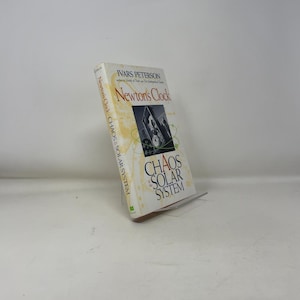 May include: A hardcover book titled "Newton's Clock" by Ivars Peterson. The cover features a photograph of a clock tower and the words "Chaos in the Solar System". The book is propped up on a clear stand.