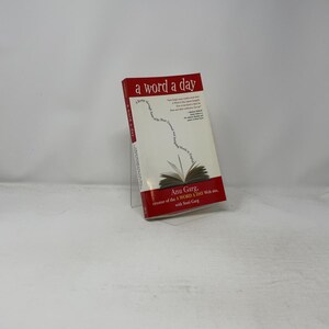 May include: A softcover book titled a word a day by Anu Garg and Stuti Garg. The cover is white with red top and bottom sections, featuring a graphic of an open book with text curving upward from the pages. The title is printed in a bold red font.