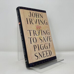 Może przedstawiać: Twarda oprawa książki Johna Irvinga zatytułowana "Trying to Save Piggy Sneed". Książka ma beżową okładkę z czarnym tekstem i ilustracją świni. Jest wyświetlana na przezroczystym akrylowym stojaku.
