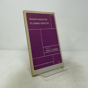 May include: A softcover book titled Behavior Modification of Learning Disabilities, edited by Robert H. Bradfield. The cover is purple with white geometric lines and white text. The spine and edges are off-white, and the price $3.95 is visible.