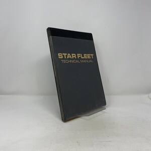 May include: A black Star Fleet Technical Manual with gold lettering. The book is slightly angled, revealing its pages and spine. The cover is a solid black color.