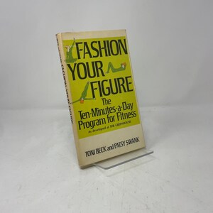 Peut inclure: Un livre vintage intitulé "Fashion Your Figure" avec une couverture jaune vif. La couverture présente des illustrations de personnes faisant de l'exercice et le texte "The Ten-Minutes-a-Day Program for Fitness". Le livre est posé sur un support transparent.
