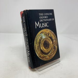 Peut inclure: Un livre à couverture rigide intitulé "The Concise Oxford Dictionary of Music" avec une couverture noire et une illustration dorée. Le livre est légèrement incliné, révélant le titre sur la tranche. Le livre est présenté sur un support transparent.