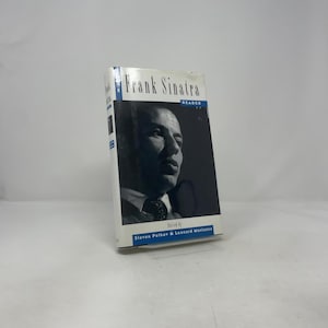 Peut inclure: Un livre intitulé "Frank Sinatra Reader" avec une image en noir et blanc de Frank Sinatra sur la couverture. Le livre a une tranche blanche et le texte "Edited by Steven Petkov & Leonard Mustazza" en bas.