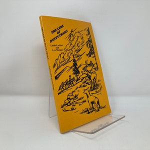 Puede incluir: Una cubierta de libro amarilla con texto negro e ilustraciones. El título es "The Lure of Dusty Trails" de Linda Locklier y Lex Mangus. La ilustración muestra un grupo de personas a caballo cabalgando por un paisaje montañoso con ciervos.