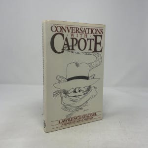 May include: A cream-coloured hardcover book titled "Conversations with Capote". The cover features a drawing of a hat, sunglasses, and a scarf. The author's name, Lawrence Grobel, and the foreword by James A. Michener are printed on the cover.