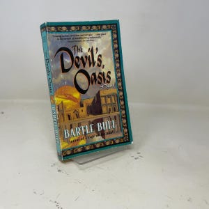 Op de afbeelding: Een paperback boek getiteld "The Devil's Oasis" van Bartle Bull. De omslag toont een woestijnscène met een grote zon en gebouwen. Het boek heeft een turquoise rand met gouden accenten en tekst.