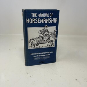 May include: A navy blue book titled "THE MANUAL OF HORSEMANSHIP" with white text. The cover features a black and white illustration of a rider on a horse jumping over an obstacle. The book is a completely revised edition by The British Horse Society and The Pony Club.