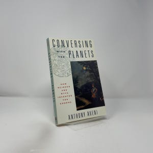 Puede incluir: Un libro de tapa dura titulado "Conversing with the Planets" de Anthony Aveni. La portada presenta una ilustración oscura de una persona en un paisaje bajo un cielo iluminado por la luna. El lomo del libro muestra el título verticalmente.