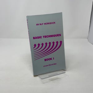 Op de afbeelding: Een grijs boek getiteld "An NLP Workbook Basic Techniques Book I" met paarse tekst en pijlgraphics. Het boek staat op een doorzichtige plastic standaard.