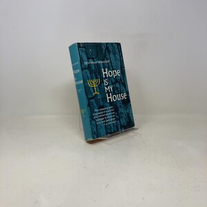 Peut inclure: Un livre à couverture turquoise intitulé "Hope Is My House" par Devorah Wigoder. Le titre est en blanc, avec un symbole de menorah jaune. La couverture présente un motif texturé en forme de pierre.
