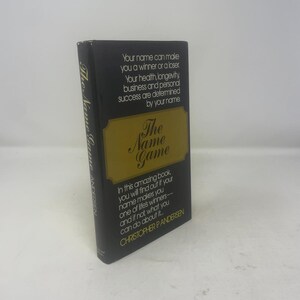 May include: A hardcover book titled "The Name Game" by Christopher P. Andersen. The book has a black cover with gold lettering and a gold rectangular design. The text on the cover discusses how a name can determine success.
