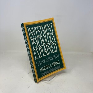 May include: A hardcover book titled "Investment Psychology Explained" by Martin J. Pring. The book has a green cover with white text and a yellow border. The book is displayed on a clear plastic stand.