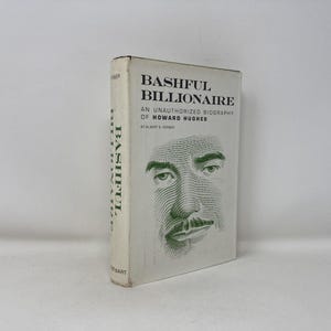 May include: A vintage hardcover book titled "Bashful Billionaire" by Albert B. Gerber. The cover features a stylized portrait of a man's face in green ink. The book is an unauthorized biography of Howard Hughes.