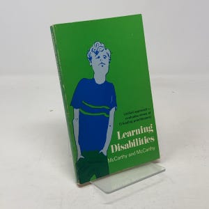 May include: A green book titled "Learning Disabilities" by McCarthy and McCarthy. The cover features a stylized illustration of a person in a blue shirt with green stripes. Text on the cover reads "Unified approach - evaluates views of 13 leading practitioners!"