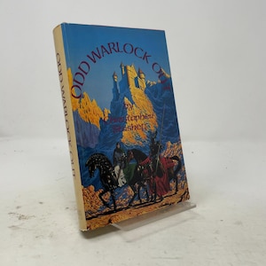 May include: A hardcover book titled Odd Warlock Out by Christopher Stasheff. The cover features a blue and yellow illustration of a castle on a mountain peak with two figures on horseback in the foreground. The spine is tan with black text.