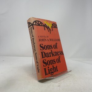May include: A vintage paperback book titled "Sons of Darkness, Sons of Light" by John A. Williams. The cover is orange with black text and a graphic of a bridge. The book is displayed on a clear acrylic stand.