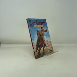 May include: A hardcover book titled "Davy Crockett at the Alamo" is displayed on a clear stand. The cover features an illustration of a man on horseback, with the Alamo in the background. The book has a blue and red cover.