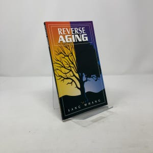 May include: A book titled "Reverse Aging" by Sang Whang. The cover features a split design with a tree silhouette. The left side has a yellow and orange gradient, while the right side is dark blue. The book is displayed on a clear stand.