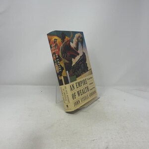 May include: A hardcover book titled "An Empire of Wealth" by John Steele Gordon. The book has a colorful cover illustration featuring a train and figures. The book is displayed on a clear plastic stand.