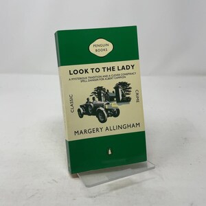 Puede incluir: Una edición de Penguin Books en verde y blanco de "Look to the Lady" de Margery Allingham. La portada del libro presenta una ilustración de coche antiguo y las palabras "Classic" y "Crime".