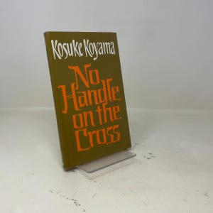 May include: A hardcover book titled "No Handle on the Cross" by Kosuke Koyama. The book has a dark olive green cover with white and orange lettering. The book is displayed on a clear plastic stand.