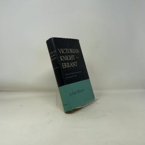 May include: A hardcover book titled "Victorian Knight Errant" by Leon Howard. The book has a black spine with white text and a teal-colored bottom half. The book is propped up on a clear stand against a white background.