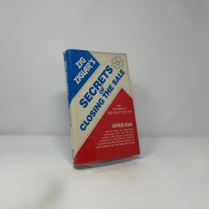 Puede incluir: Un libro de tapa dura titulado "Secrets of Closing the Sale" de Zig Ziglar. El libro tiene una portada azul, blanca y roja con el título en letras grandes y en negrita. El libro promete más de 100 cierres específicos para vendedores.