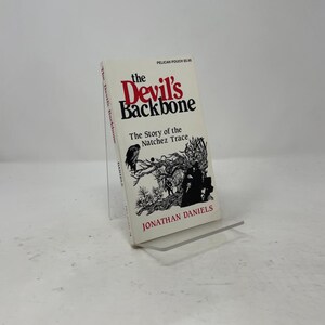 Könnte beinhalten: Ein Taschenbuch mit dem Titel The Devil's Backbone: The Story of the Natchez Trace von Jonathan Daniels. Der weiße Einband zeigt schwarzen Text, eine schwarz-weiße Illustration eines Vogels und Figuren im Wald sowie das Pelican Pouch Logo.