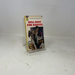 Può includere: Un libro tascabile vintage intitolato Hell-Bent for Danger di Walt Grove. La copertina presenta uno sfondo color crema con testo rosso, un'illustrazione centrale di una persona in una stanza buia e una mano che tiene un bicchiere. Il prezzo 25 cent è stampato.