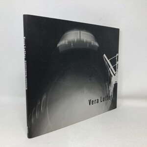 Puede incluir: Portada de libro de fotografía en blanco y negro con el título "Vera Lutter" y una foto de un objeto grande, oscuro y circular con una forma rectangular clara en la parte superior.