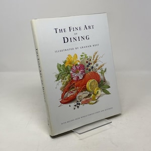 May include: A hardcover book titled "The Fine Art of Dining" with illustrations by Graham Rust. The cover features a vibrant still life of a lobster, lemons, flowers, and other food items. The book is propped up on a clear stand.