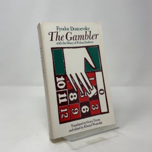 May include: A paperback book titled "The Gambler" by Fyodor Dostoevsky. The cover features a hand over a roulette wheel design with numbers in red and white. The book is translated by Victor Terras and edited by Edward Wasiolek.