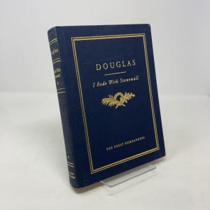 Può includere: Un libro con copertina rigida blu scuro intitolato "DOUGLAS I Rode With Stonewall" della serie "The Great Commanders". Il libro presenta scritte dorate e un elemento decorativo dorato. Il libro è esposto su un supporto trasparente.
