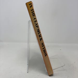 May include: A vintage book titled "In the Clap Shack / Styron" with a tan cover and black text. The book is displayed on a clear acrylic stand, showcasing its spine. The book's edges show signs of wear.