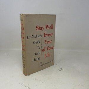 Könnte beinhalten: Ein Vintage-Buch mit dem Titel "Stay Well: Dr. Molner's Every Year Guide To Your Total Health of Your Life" wird angezeigt. Das Buch hat einen beigefarbenen Einband mit rotem und schwarzem Text. Das Buch steht auf einer weißen Oberfläche.